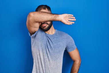 Middle east man with beard standing over blue background covering eyes with arm, looking serious and sad. sightless, hiding and rejection concept 