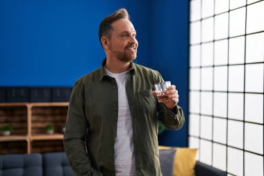 Genç beyaz adam evde dikilip bir bardak viski içiyor.