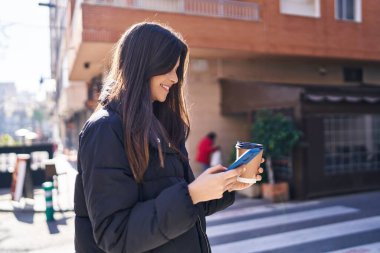 Genç, güzel, İspanyol bir kadın sokakta akıllı telefon kullanarak gülümsüyor.