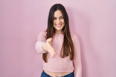 Young brunette woman standing over pink background smiling cheerful offering palm hand giving assistance and acceptance. 