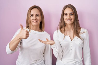 Orta yaşlı anne ve küçük kızı pembe arka planda durup avuç içini gösteriyor ve baş parmaklarıyla iyi bir jest yapıyor, mutlu ve neşeli gülümsüyor. 
