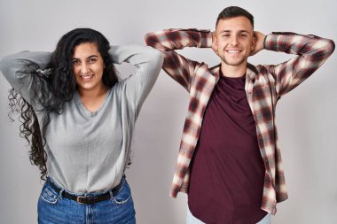 Beyaz arka planda duran genç İspanyol çift rahatlıyor ve esniyor, kolları ve elleri başının arkasında ve boynunun arkasında mutlu bir şekilde gülümsüyor. 