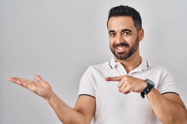Young hispanic man with beard wearing casual clothes over white background amazed and smiling to the camera while presenting with hand and pointing with finger. 