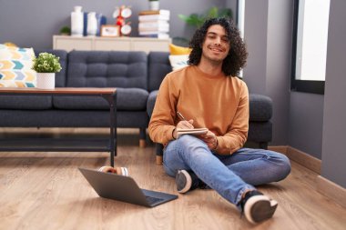 Young latin man using laptop writing on notebook at home