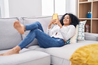 Evdeki kanepede uzanmış akıllı telefon kullanan genç İspanyol kadın.