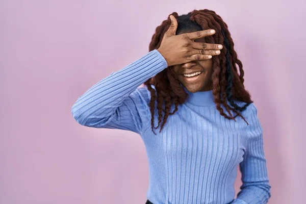 ピンクの背景の上に立つアフリカの女性は笑顔で 驚きのために目を覆う顔で手で笑っています 視覚障害の概念 — ストック写真