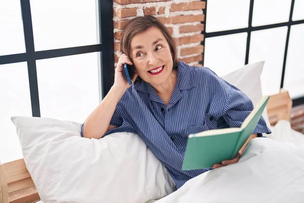 Middle age woman talking on smartphone reading book at bedroom