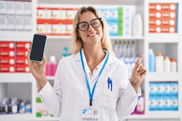 Young blonde woman working at pharmacy drugstore showing smartphone screen smiling happy pointing with hand and finger to the side