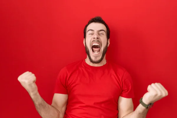 カジュアルな赤いTシャツを着た若いヒスパニック系の男性は 腕を上げて目を閉じて成功に驚いて驚いています 勝者のコンセプト — ストック写真