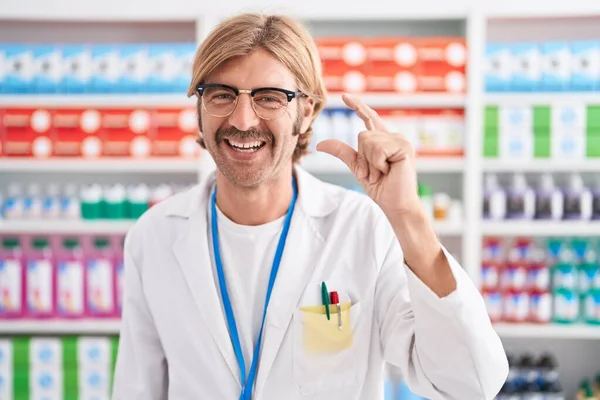 Caucasian man with mustache working at pharmacy drugstore smiling and confident gesturing with hand doing small size sign with fingers looking and the camera. measure concept.