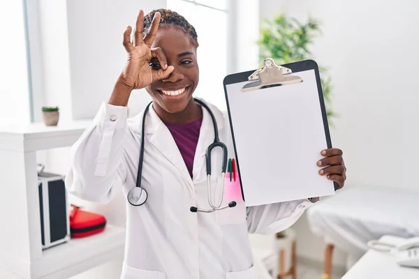 Beautiful black doctor woman holding clipboard smiling happy doing ok sign with hand on eye looking through fingers