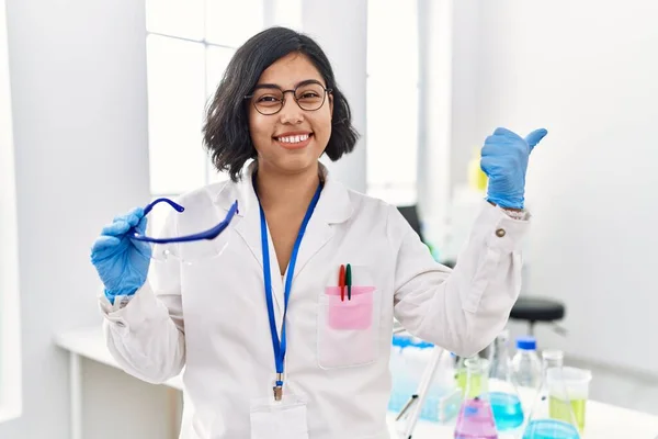 Young hispanic woman working at scientist laboratory holding safety glasses pointing thumb up to the side smiling happy with open mouth