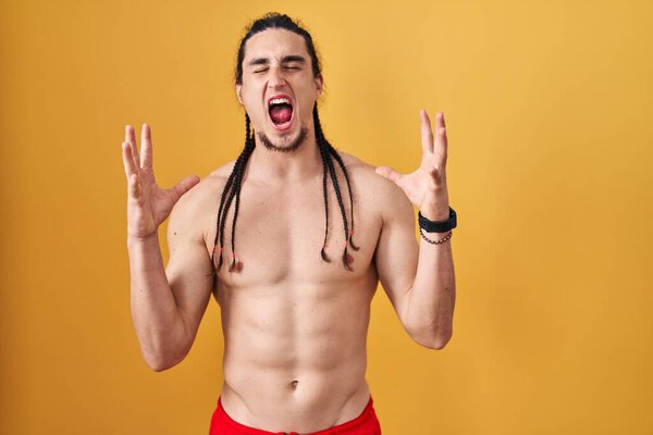 Hispanic man with long hair standing shirtless over yellow background celebrating mad and crazy for success with arms raised and closed eyes screaming excited. winner concept 