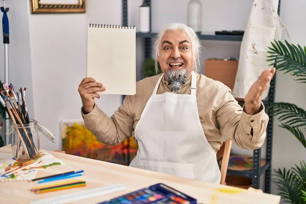 白髪交じりの中年男性がアートスタジオに座り 幸せな笑顔と受賞者の表情でノートを持っています — ストック写真
