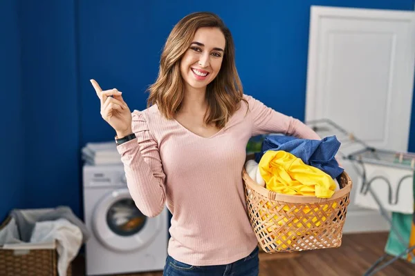 Young woman holding laundry basket with a big smile on face, pointing with hand finger to the side looking at the camera.