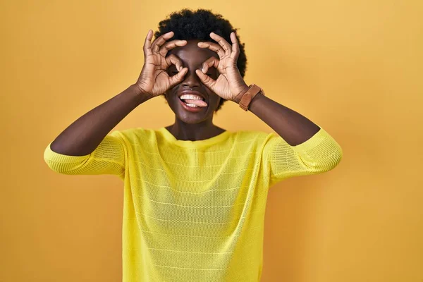 黄色のスタジオの上に立っているアフリカの若い女性は 双眼鏡が舌を突き出すように 目は指を通して見て をジェスチャーを行います クレイジーな表現 — ストック写真