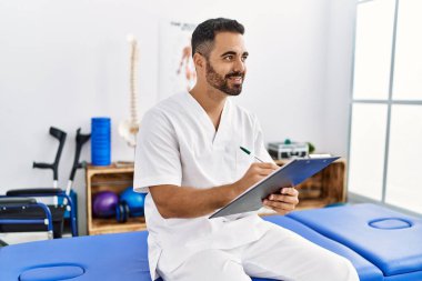 Fizyoterapist üniforması giyen genç İspanyol adam klinikteki panoya yazı yazıyor.