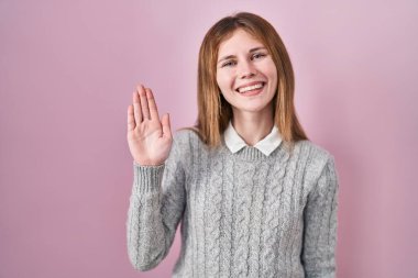Beautiful woman standing over pink background waiving saying hello happy and smiling, friendly welcome gesture 