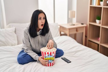 Hispanic woman eating popcorn watching a movie on the bed in shock face, looking skeptical and sarcastic, surprised with open mouth 