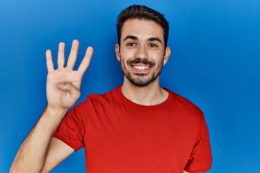 Young hispanic man with beard wearing red t shirt over blue background showing and pointing up with fingers number four while smiling confident and happy. 