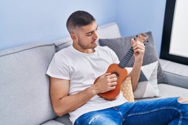 Young hispanic man playing ukelele sitting on sofa at home