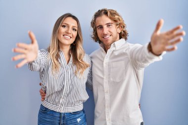 Mavi arka planda duran genç çift kameraya bakıp kucaklaşmak için kollarını açmış gülümsüyor. Mutluluğu kucaklayan neşeli bir ifade.. 