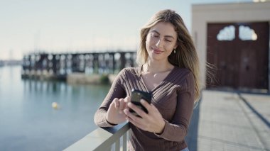 Young blonde woman smiling confident using smartphone at seaside