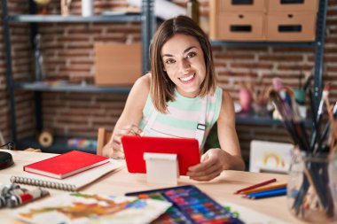 Young beautiful hispanic woman artist using touchpad sitting on table at art studio