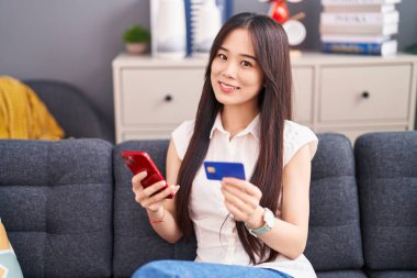 Akıllı telefon ve kredi kartı kullanan Çinli genç bir kadın evdeki kanepede oturuyor.