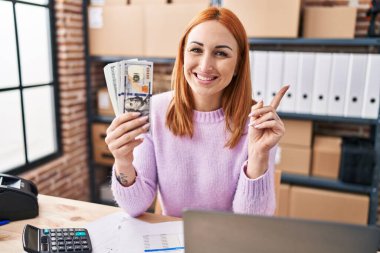 Young woman working at small business ecommerce holding money smiling happy pointing with hand and finger to the side 