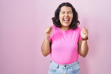 Pembe arka planda duran genç İspanyol kadın başarı için heyecanlı kolları havada ve gözleri kapalı zaferi kutlarken gülümsüyor. Kazanan konsept. 