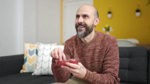 Young bald man eating raspberries sitting on sofa at home