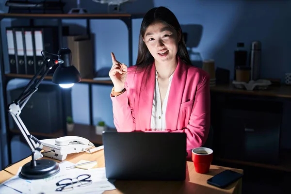 Chinese young woman working at the office at night with a big smile on face, pointing with hand and finger to the side looking at the camera.