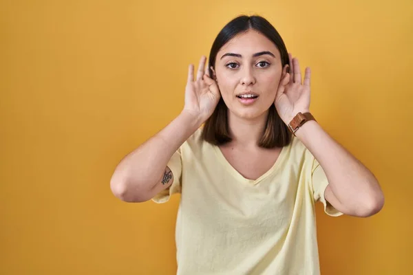 西班牙裔女孩穿着黄色背景的休闲装 试图听到双手放在耳朵上的手势 好奇地闲聊 听力问题 — 图库照片