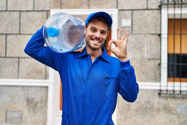 Young hispanic man holding a gallon bottle of water for delivery doing ok sign with fingers, smiling friendly gesturing excellent symbol