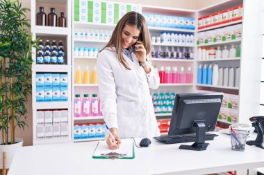 Young beautiful hispanic woman pharmacist talking on smartphone writing on document at pharmacy