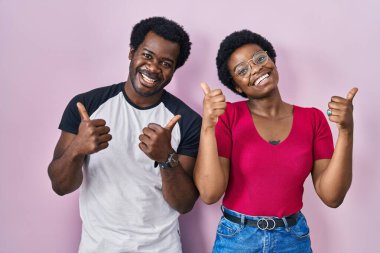 Genç Afro-Amerikan çifti pembe arka plan başarısı tabelasının üzerinde duruyorlar. Elleriyle olumlu bir jest yapıyorlar. Gülümsüyorlar ve mutlular. Neşeli ifade ve kazanan jest. 