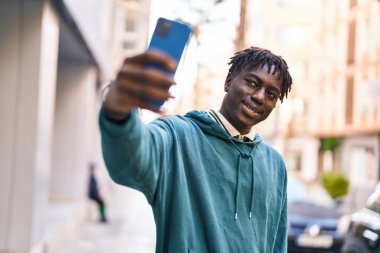 Afro-Amerikalı adam gülümsüyor. Kendine güveni tam. Sokakta akıllı telefondan selfie çekiyor.