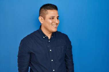 Mavi arka planda duran İspanyol genç adam yüzünde gülümseme ve doğal ifadeyle yan yana bakıyor. Kendinden emin gülen. 