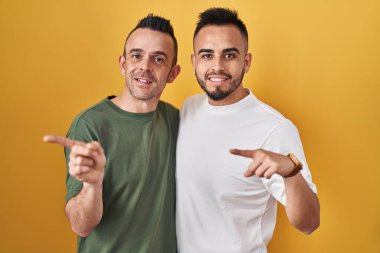 Sarı arka planda dikilen eşcinsel çift, elleri ve başparmakları havada, kendinden emin bir şekilde sırıtıyor. 