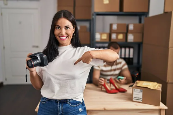 Young hispanic woman holding camera working at small business ecommerce pointing finger to one self smiling happy and proud