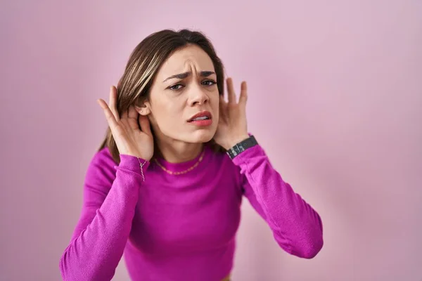 西班牙裔妇女站在粉红的背景上试图听到双手放在耳朵上的手势 好奇地闲聊 听力问题 — 图库照片