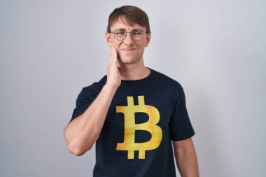 Bitcoin t-shirt giyen beyaz sarışın adam diş ağrısı ya da diş hastalığından dolayı ağıza acı dolu bir ifadeyle dokunuyor. dişçi 