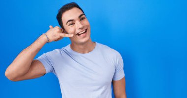 Young hispanic man standing over blue background smiling doing phone gesture with hand and fingers like talking on the telephone. communicating concepts. 