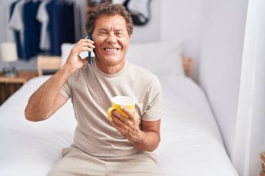 Orta yaşlı bir adam akıllı telefondan konuşuyor. Yatak odasında kahve içiyor.