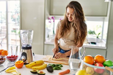 Genç, güzel İspanyol kadın mutfakta muz kesen blenderlı sebze suyu hazırlıyor.