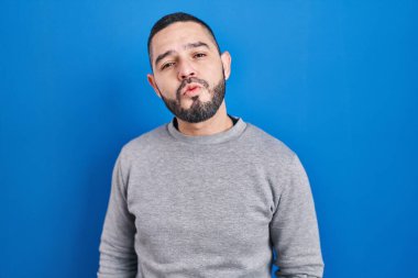 Latin asıllı bir adam mavi arka planda dikilmiş kameraya bakıyor ve güzel ve seksi bir şekilde yayına öpücük yolluyor. aşk ifadesi. 