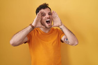 Young hispanic man standing over yellow background smiling cheerful playing peek a boo with hands showing face. surprised and exited 