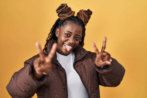 African woman with braided hair standing over yellow background smiling with tongue out showing fingers of both hands doing victory sign. number two.