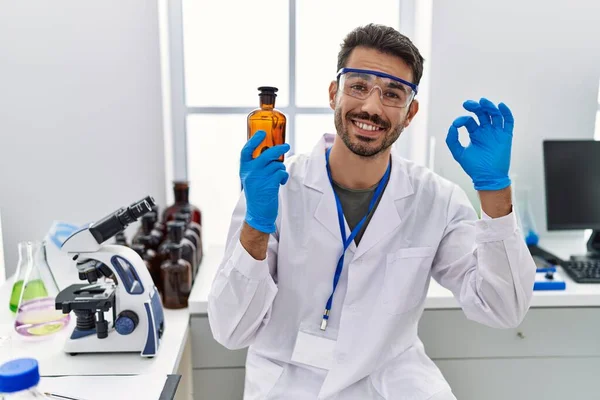 Joven Hombre Hispano Trabajando Laboratorio Científico Sosteniendo Botella Haciendo Signo — Foto de Stock